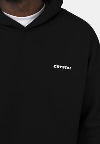Mustapaita, joka on valmistettu puuvillasta ja jossa on valkoinen "CRYSTAL"-logo vasemmassa rinnassa. Huoodattu muotoilu, jossa on pehmeä tekstuuri.