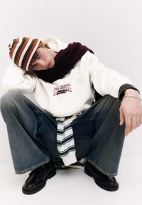 Witte hoodie met "CLASSIC" opdruk, blauwe wijde jeans, gestreepte pet in groen en oranje, gestreept shirt om de taille gestrikt, zwarte schoenen.