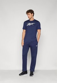 Marineblaues Sportoutfit mit kurzärmeligem Shirt und weißem Logo sowie passenden Hosen aus glattem Stoff, getragen mit schwarzen Sneakern.