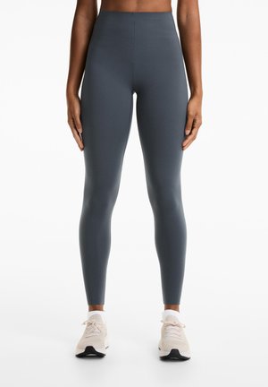 Grijze high-waisted leggings met een gladde, rekbare textuur, strakke pasvorm en geen zichtbare naden. Een paar beige sportschoenen worden gedragen.