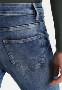 Siniset farkut, joissa on haalistunut pesu, taustatasku, vyölenkit ja näkyvät ompeluyksityiskohdat. Kankaan tekstuuri on rento ja suunnittelu.