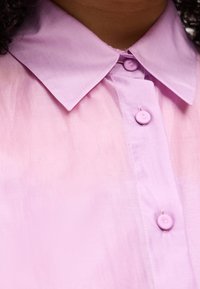 Εγγύς πλάνο ενός ατόμου που φοράει ένα πουκάμισο ανοιχτού μωβ χρώματος με κουμπιά και μυτερό γιακά σε ταιριαστή μωβ απόχρωση.