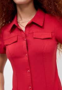 Chemise rouge à manches courtes avec une coupe ajustée, dotée d'une patte de boutonnage, de deux poches poitrine et d'une texture lisse.