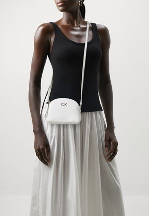 Vit läder crossbody-väska med en slät textur, rund form och CK-logotyp i silver. Bärs med en svart linne och vit kjol.