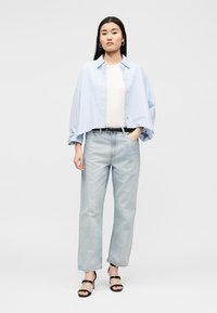 Γυναίκα που φοράει ανοιχτό μπλε πουκάμισο oversized πάνω από λευκή μπλούζα, ανοιχτόχρωμο τζιν, μαύρα πέδιλα με τακούνι, που στέκεται σε απλό φόντο.