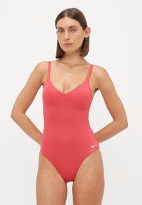 Jednodílné plavky v jasně korálové barvě, vyrobené z hladkého materiálu. Mají V-výstřih, tenké ramínka a malé bílé logo Nike na bocích.