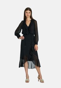 Vestido negro con escote en V rizado y mangas largas y transparentes, que presenta una falda fluida con dobladillo asimétrico. Combinado con tacones con tiras.
