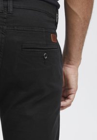 Czarne spodnie z tylną kieszenią zapinaną na guzik oraz skórzaną metką. Tkanina ma gładką fakturę i klasyczny design.