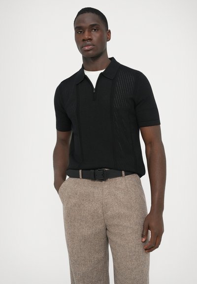 Polo preta de manga curta com gola com fecho de correr, apresentando painéis texturizados; combinada com calças bege texturizadas e um cinto preto.