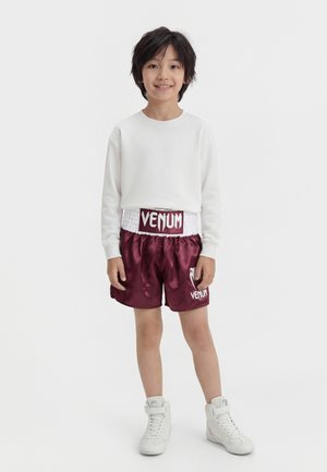 Otrok s temnimi lasmi, oblečen v belo dolgo majico, bordo boksarske hlače Venum in bele superge z visokim zgornjim delom, stoji in se smehlja.