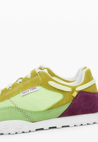 Semišové a textilní tenisky v zelené, žluté a fialové barvě. Mají bílou podrážku, prošívané detaily a štítek s nápisem "Od roku 1984."