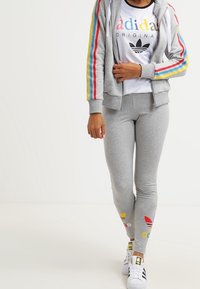 Grå zip-up hoodie med flerfärgade sidosträck, tillsammans med grå leggings med logomönster och accentuerad med vita sneakers.