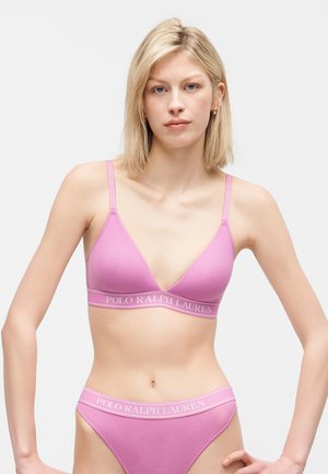 Γυναίκα με ξανθά μαλλιά που φοράει ροζ σουτιέν Polo Ralph Lauren και ταιριαστά εσώρουχα μπροστά σε απλό λευκό φόντο.