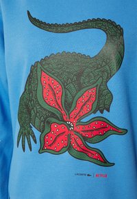 Blå bomullströja med en grön reptildesign som är sammanflätad med en röd blomma. Inkluderar svarta konturer samt Lacoste- och Netflix-logotyper.