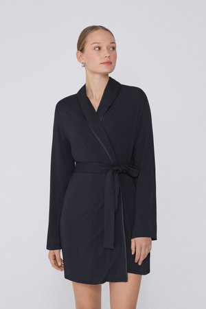 Jeune femme aux cheveux attachés portant une robe noire longue jusqu'aux genoux, à manches longues et ceinture nouée à la taille, regardant vers sa droite.