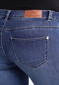 Jeans de mezclilla azul oscuro con una etiqueta de marca de cuero en la cinturilla y un bolsillo trasero con un detalle de remache plateado.