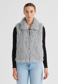 Faux-Fur-Weste in Hellgrau mit Kragen, vertikalen Streifen und einem vorderen Reißverschluss. Getragen über einem schwarzen Langarmoberteil und Jeans.