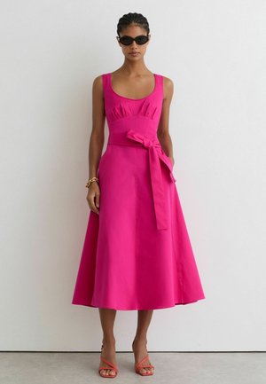 Žena v bezrukávových svetloružových midi šatách s opaskom v páse, tmavých slnečných okuliaroch, zlatom náramku a koralových otvorených lodičkách stojí pri bielej stene.