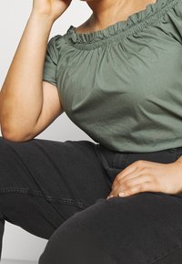 Top verde oliva con escote bardot, con cuello elástico fruncido, combinado con pantalones negros de cintura alta. Tejido suave, corte holgado.