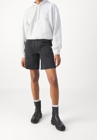 Šedá mikina s kapucí a přední kapsou, v kombinaci s černými denimovými kraťasy. Černé kotníkové boty s tlustými podrážkami a bílé žebrovan�é ponožky. Jednoduché pozadí.