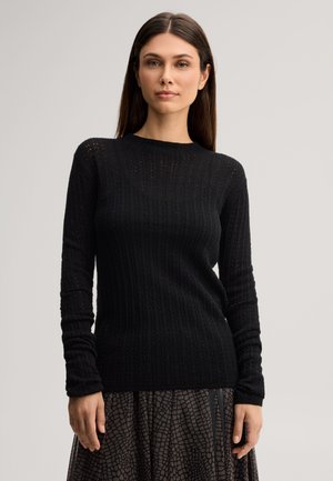 Jeune femme aux cheveux bruns raides portant un pull en maille noir à manches longues et une jupe à motifs, debout devant un fond uni.