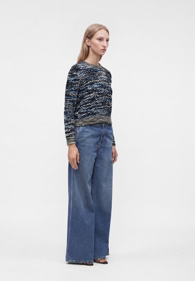 Femme portant un pull bleu à motif, un jean bleu à jambes larges et des chaussures noires à bouts ouverts, debout devant un fond blanc uni.