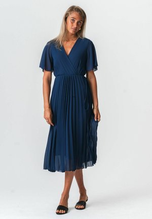 Žena s dlhými blond vlasmi, ktorá má na sebe námornícky modré plisované midi šaty s roztiahnutými rukávmi a čierne sandále s otvorenou špičkou, stojaca na bielom pozadí.