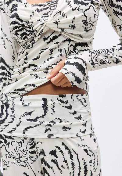 Κοντινό πλάνο κορμού με λευκό τοπ μακρυμάνικο και φούστα με αφηρημένο μαύρο μοτίβο τίγρης, με το χέρι να σηκώνει ελαφρώς το τοπ για να αποκαλύψει την κοιλιά.