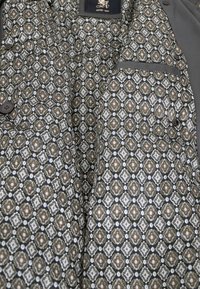 Braun-weißer gemusterter Stoff mit geometrischen Designs, der ein äußeres Kleidungsstück auskleidet. Besitzt Knöpfe und eine Seitentasche auf der linken Seite.