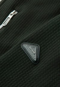 Svart texturerad tyg med en triangulär logolapp med texten "quintessence" och en silverdragkedjedetalj.