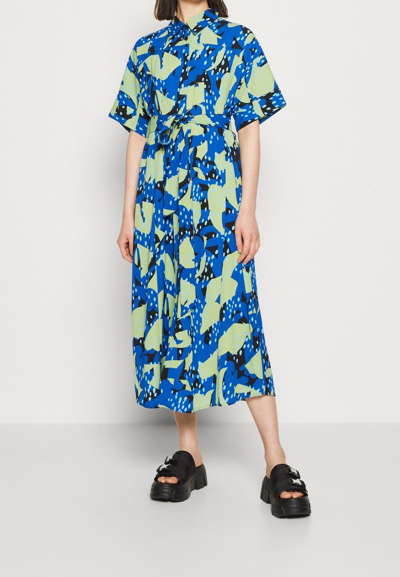 Blau- und grün gemustertes Kleid mit kurzen Ärmeln, tailliertem Bund und Midi-Länge. Kombiniert mit klobigen schwarzen Plateau-Sandalen.