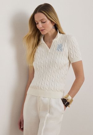 Kobieta ubrana w biały, krótki sweter z okrągłym dekoltem i warkoczowym splotem, białe spodnie oraz złote i bursztynowe bransoletki, stojąca na tle gładkiej ściany.