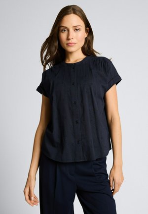 Junge Frau mit langen braunen Haaren, die eine kurzärmlige schwarze Bluse mit Knöpfen und schwarze Hosen trägt, vor einem einfarbigen Hintergrund.