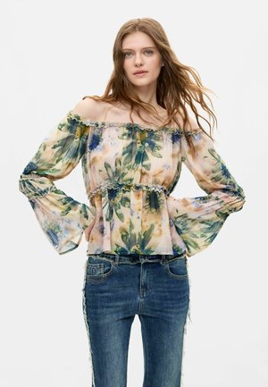 Frau trägt eine schulterfreie, langärmelige Blumenbluse mit Rüschen und blaue Jeans und steht vor einem schlichten, hellen Hintergrund.