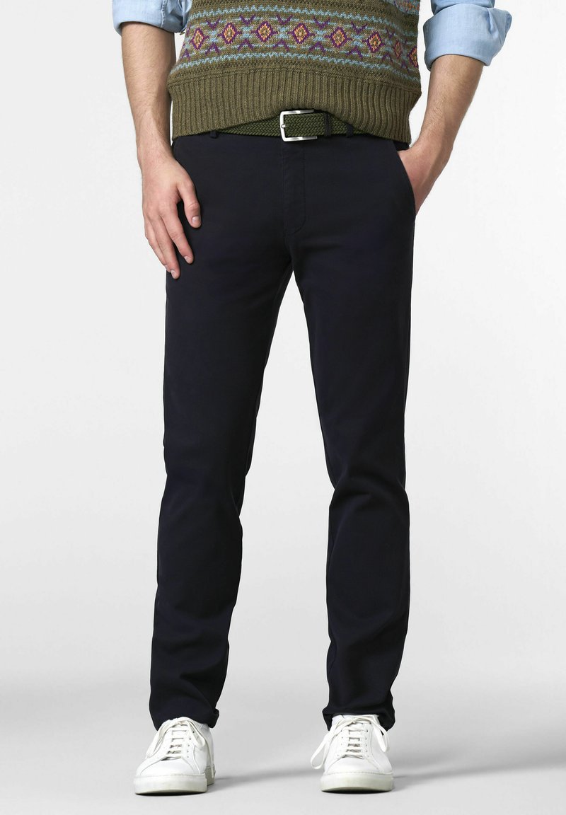 Homme portant une chemise bleue retroussée, un gilet en laine olive à motifs, un pantalon sombre avec une ceinture verte et des baskets blanches, se tenant avec les mains en partie dans les poches.