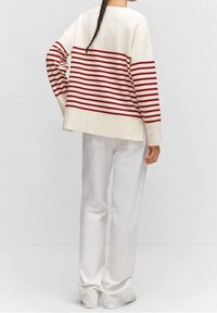 Henkilö, jolla on letitetyt hiukset, pukeutuneena kermaiseen neuleeseen, jossa on punaisia vaakaraitoja, ja valkoisiin pantsareihin, seisoo käsi lantiolla ja katsoo poispäin.