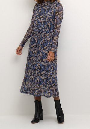 Kvinde iført en langærmet, blå og beige blomsterprint midi-kjole kombineret med sorte ankelstøvler med blokhæl mod en ensfarvet baggrund.