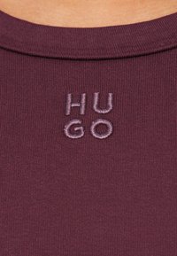 Camisa de algodão borgonha com decote redondo, apresentando o logótipo "HUGO" bordado em fio lilás claro perto da gola. Tecido texturizado.