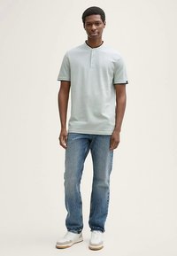 Šviesiai žalia trumpomis rankovėmis polo marškinėliai su trimis mygtukais ir juoda apykakle, derinami su mėlynais džinsais ir baltais sportbačiais.