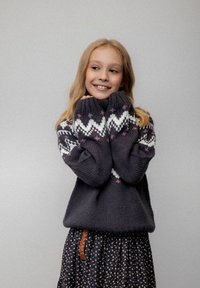 Uśmiechnięta młoda dziewczyna z długimi blond włosami, ubrana w ciemny sweter z dzianiny w białe i różowe wzory oraz ciemną spódnicę w kwiaty z plecionym paskiem.