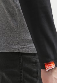 En person som bär en svart långärmad tröja med en röd Superdry-etikett på ärmen, kombinerad med en grå topp och svarta byxor.