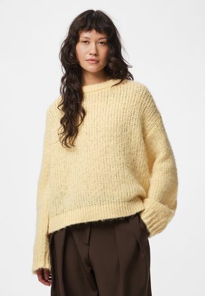 Femme aux longs cheveux ondulés portant un pull en maille jaune ample et un pantalon large marron foncé sur un fond blanc uni.