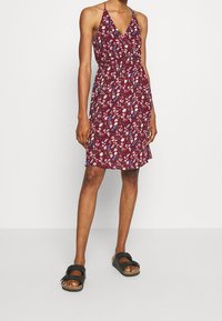 Burgundské květinové šaty s výstřihem do V, zavazováním za krk a zúženým pasem. Mají splývavou sukni a jsou nošeny s černými sandály.