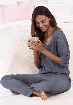 Grau gestricktes Lounge-Set mit einem V-Ausschnitt-Oberteil und lockeren Hosen. Sitzt auf einem Bett mit strukturiertem cremefarbenem Bettzeug und hält eine Tasse.