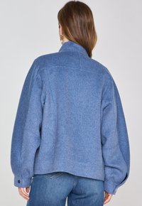 Giacca blu in misto lana con una vestibilità leggermente oversize, colletto alto e polsini con bottoni. Texture liscia, design casual e semplici dettagli di cucitura.