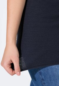 Maglia a maglia blu navy con un motivo strutturato, caratterizzata da un orlo con una leggera lavorazione a rete. La mano afferra il bordo inferiore, abbinata a jeans di denim.