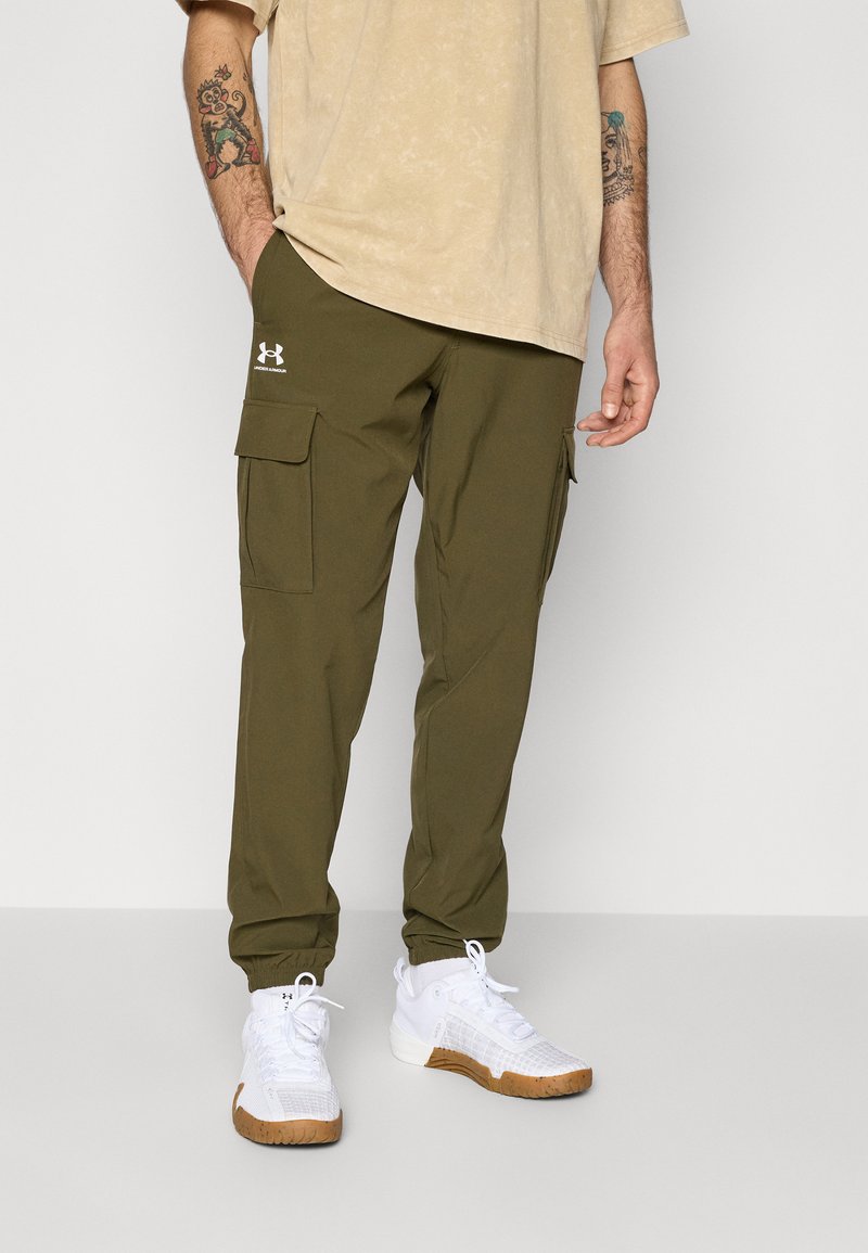 Olive green cargo pants with two side pockets, tapered cuffs, and Under Armour logo in white. Worn with a beige T-shirt and white sneakers.