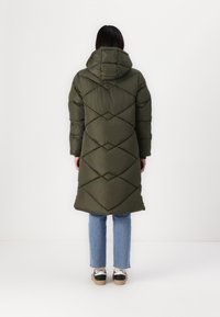 Manteau matelassé long vert olive avec un motif à losanges, capuche et manches, associé à un jean bleu et des baskets noir et blanc.