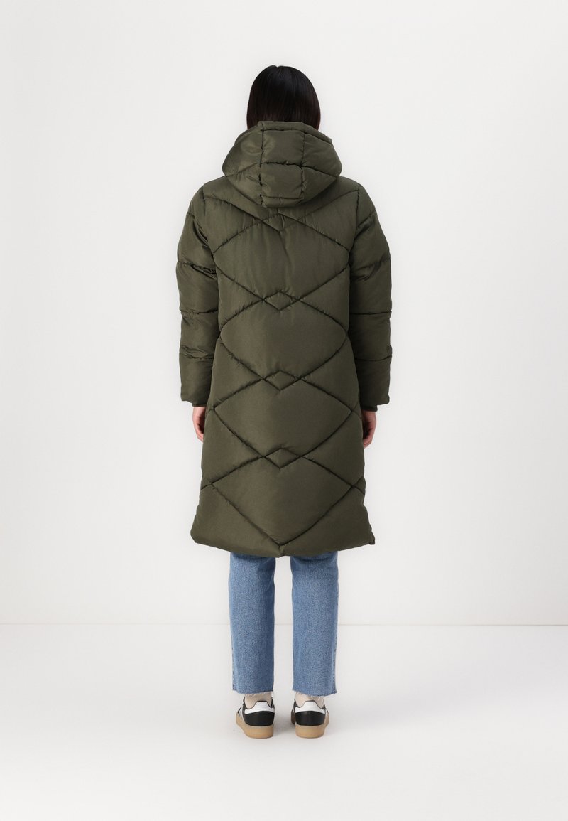 Langer olivgrüner Puffer-Mantel mit einem Rautenmuster, Kapuze und Ärmeln, kombiniert mit blauen Jeans und schwarz-weißen Sneakers.