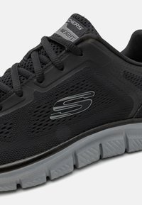 Schwarzer Sportschuh mit Mesh-Obermaterial, strukturierten Details, grauen Akzenten und einer gepolsterten Sohle mit einem gemusterten Profil. Logo an der Seite angebracht.